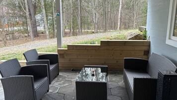 A covered patio area with wicker furniture and a stone floor, overlooking a wooded area.
