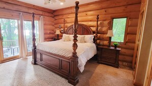 A log cabin bedroom with a four-poster bed, a nightstand, a lamp, and a view of the outdoors through a sliding glass door. - Beautiful Chalet on the Lake! (Carleton Place)
