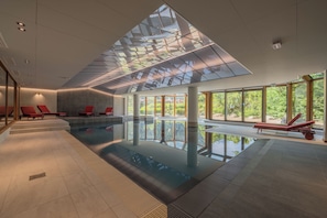 A modern indoor swimming pool with clear glass ceiling, red lounge chairs, and large windows.
