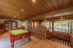 A wooden pool table in a room with a wooden ceiling and a balcony with a railing.