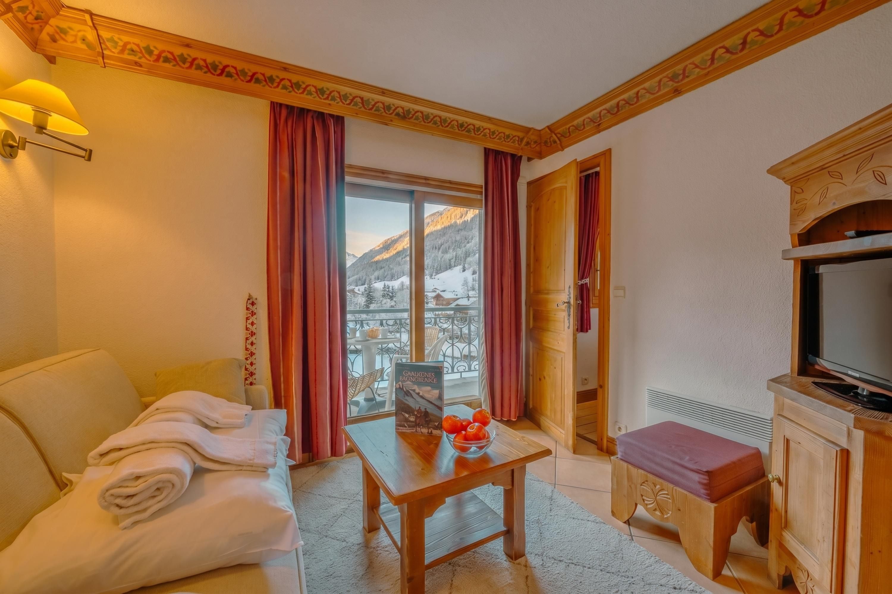 Une chambre d’hôtel avec un canapé, une table avec un livre et des fruits, une télévision et un balcon offrant une vue sur la montagne.