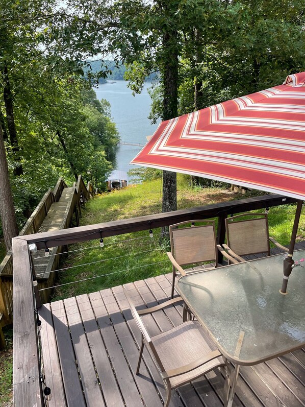 A wooden deck with a glass table and chairs, overlooking a lake.