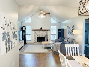 Cozy living space with a stone fireplace, high ceiling, and large windows offering a view of the outdoors.