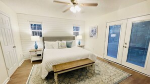 Un dormitorio con cama, mesitas de noche, ventilador de techo y un amplio ventanal con persianas.