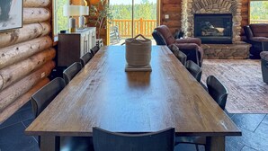 Interior de uma cabana de toras com uma longa mesa de jantar, lareira de pedra e vista para a floresta.