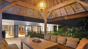 An outdoor seating area with a wooden table and patterned cushions, a wooden ceiling structure, and a view of the interior space.