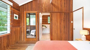 Ein Schlafzimmer mit Holzwänden, einem Bett, einem Nachttisch, einer Lampe und Blick auf ein Badezimmer und eine Küche durch eine offene Tür.