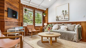 Ein gemütliches Wohnzimmer mit Holzvertäfelung, einem runden Holztisch, einer Couch mit Kissen, einem an der Wand montierten Fernseher und einem Fenster mit Blick auf Grünflächen.