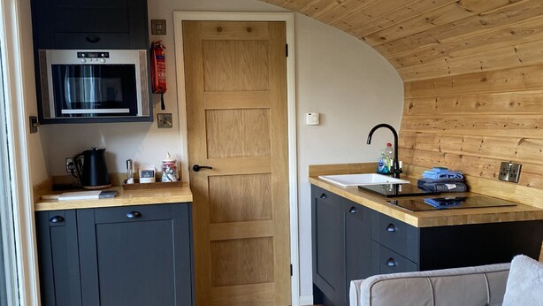 A compact kitchen with a microwave, sink, and dark cabinets. There's a wooden door leading to another room and a sofa in the corner. - Meadow View  - (Bottom Cabin)  - sleeps 2 guests  in 1 bedroom (Foolow)