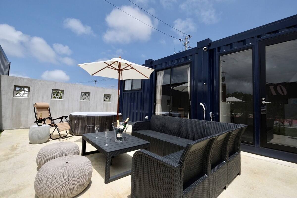 An outdoor patio with a dining table, chairs, and a large umbrella. There is a hot tub and a sofa in the background.
