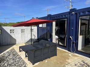 A compact outdoor seating area with a black wicker sofa and a red umbrella, adjacent to a blue container-style building with a sliding glass door leading to a bedroom. - SORARU MIYAKOJIMA (Miyakojima)