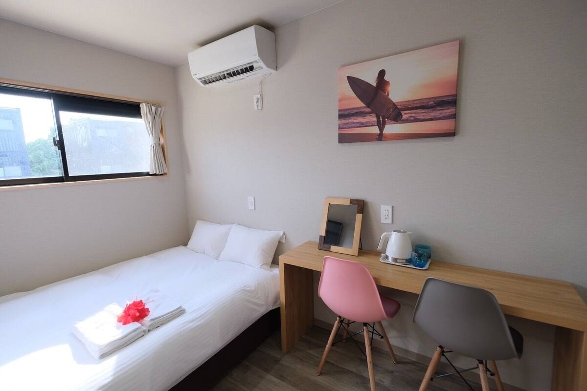 A single bed with white bedding, a desk with a mirror, a pink chair, and a picture of a surfer on the wall.