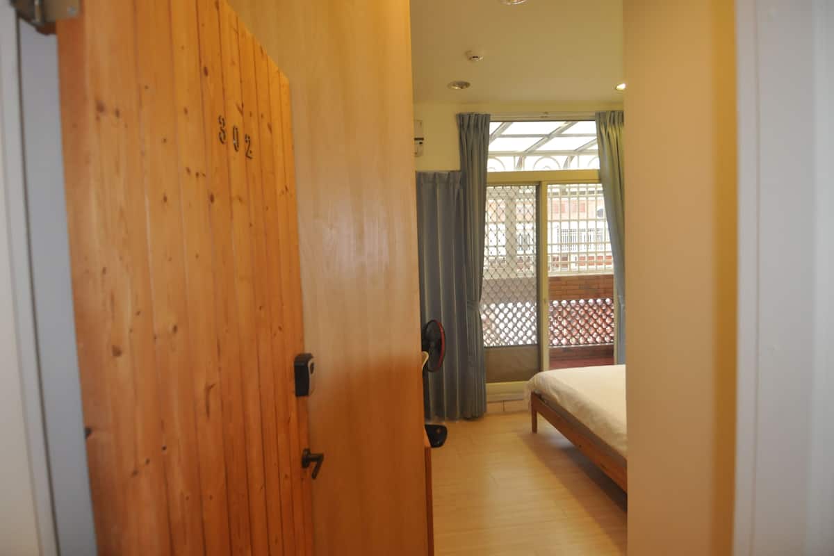 Un dormitorio con puerta de madera, cama, ventana con cortinas y vista a un balcón.