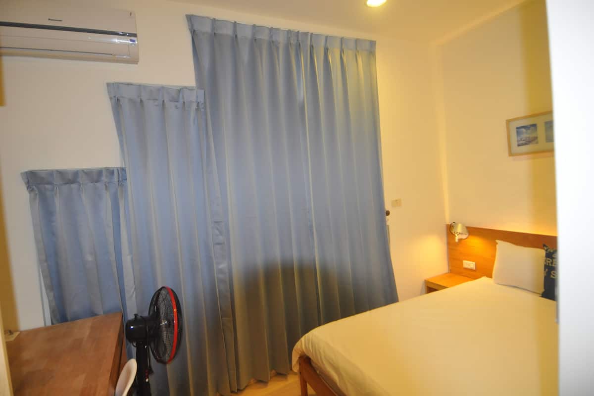 Una habitación de hotel con una cama, una mesita de noche, una lámpara, un ventilador y cortinas.