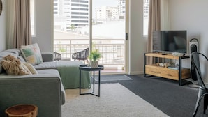 A modern living room with a sofa, a TV on a wooden stand, and a balcony with a city view. - Inner City One Bedroom Apartment with Pool View 22 (Cairns)