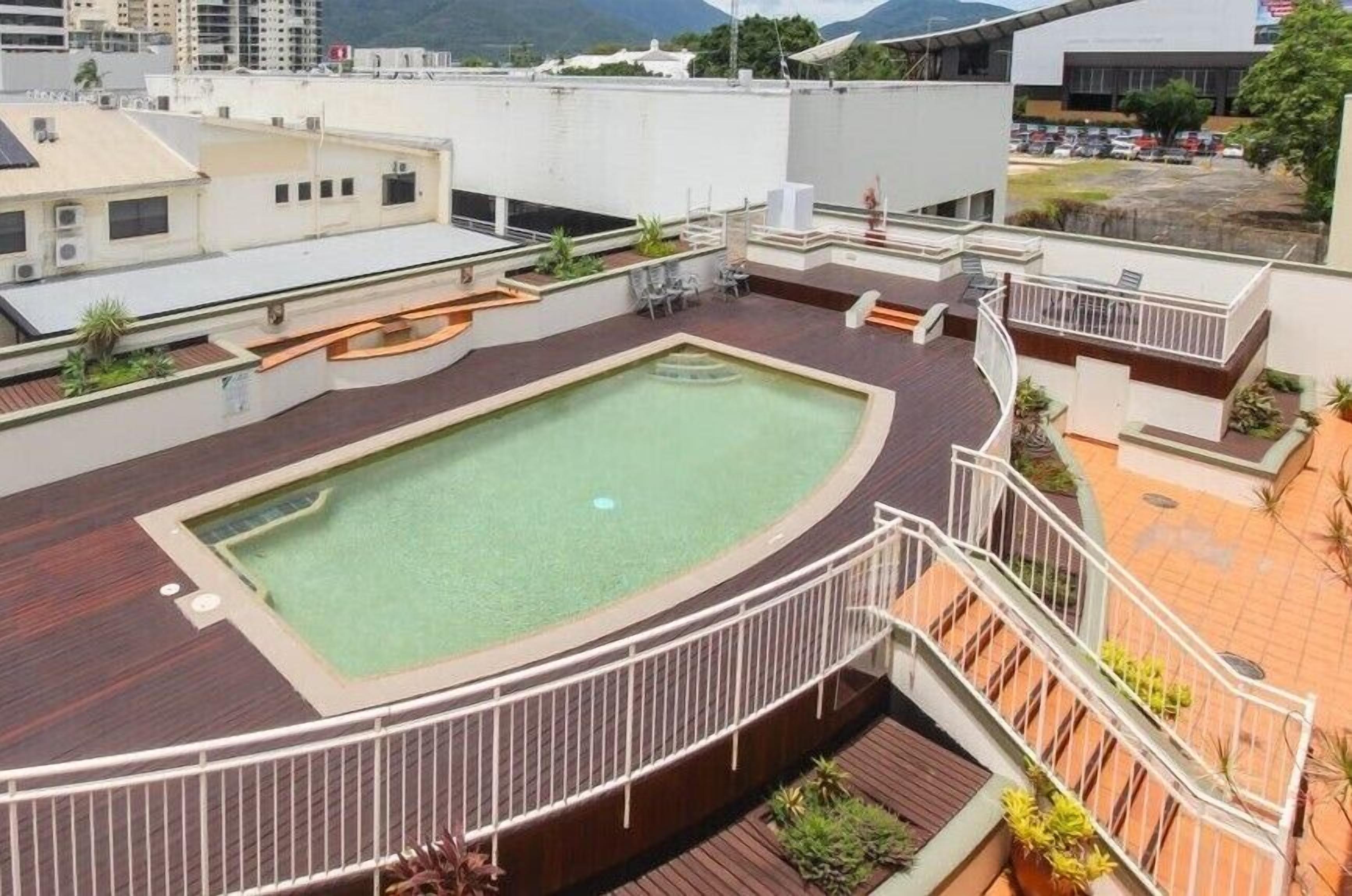 A rooftop pool area with a circular pool, surrounded by wooden decking and railings, with buildings and a parking lot in the background.