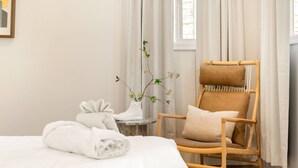 A neatly made bed with white linens, a wooden chair with cushions, a small table with a vase of flowers, and a window with curtains. - Voted Guest Favorite! Get Wow'd! Stellar Views Hollywood Hills (Los Angeles)