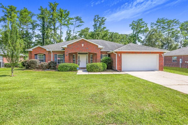 A two-story brick house with a white garage door, surrounded by a well-maintained lawn and trees. - Spacious 4 bedroom house by OWA & Tanger Outlets, 15 mins from Beaches (Foley)