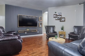 A modern living room with a large flat-screen TV mounted on a grey wall, leather sofas, a wooden coffee table, and a small side table with a plant.