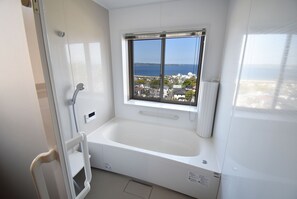 A modern bathroom with a bathtub, a window with a view of the sea, and a white shower area. - Bep one (Beppu)