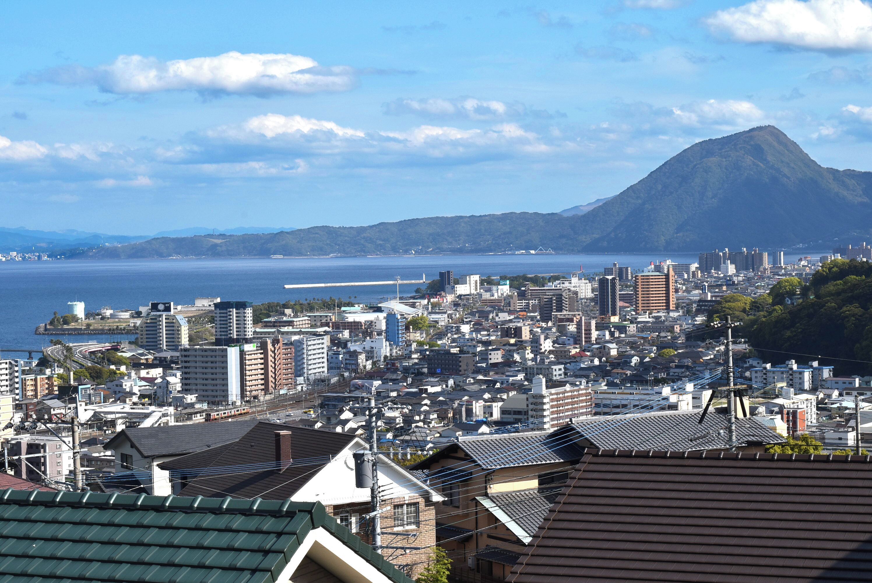 A coastal cityscape with a prominent mountain, dense urban buildings, and a clear view of the sea.