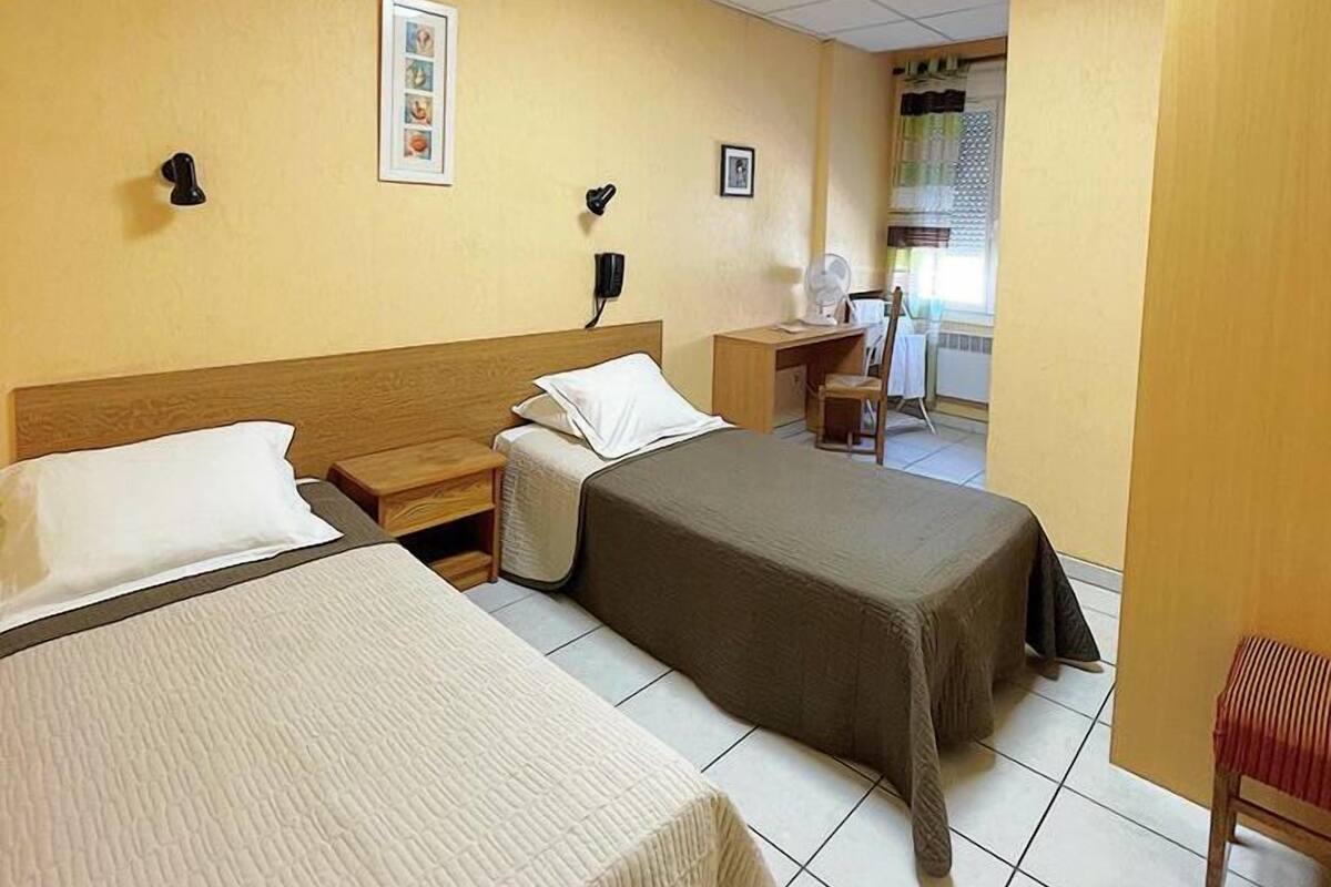 Une chambre d’hôtel avec deux lits, un bureau, une chaise et une fenêtre avec des rideaux.