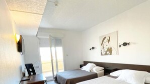 Une chambre d’hôtel avec deux lits simples, un bureau et une chaise. Il y a une fenêtre avec des stores, une horloge murale et un tableau encadré.