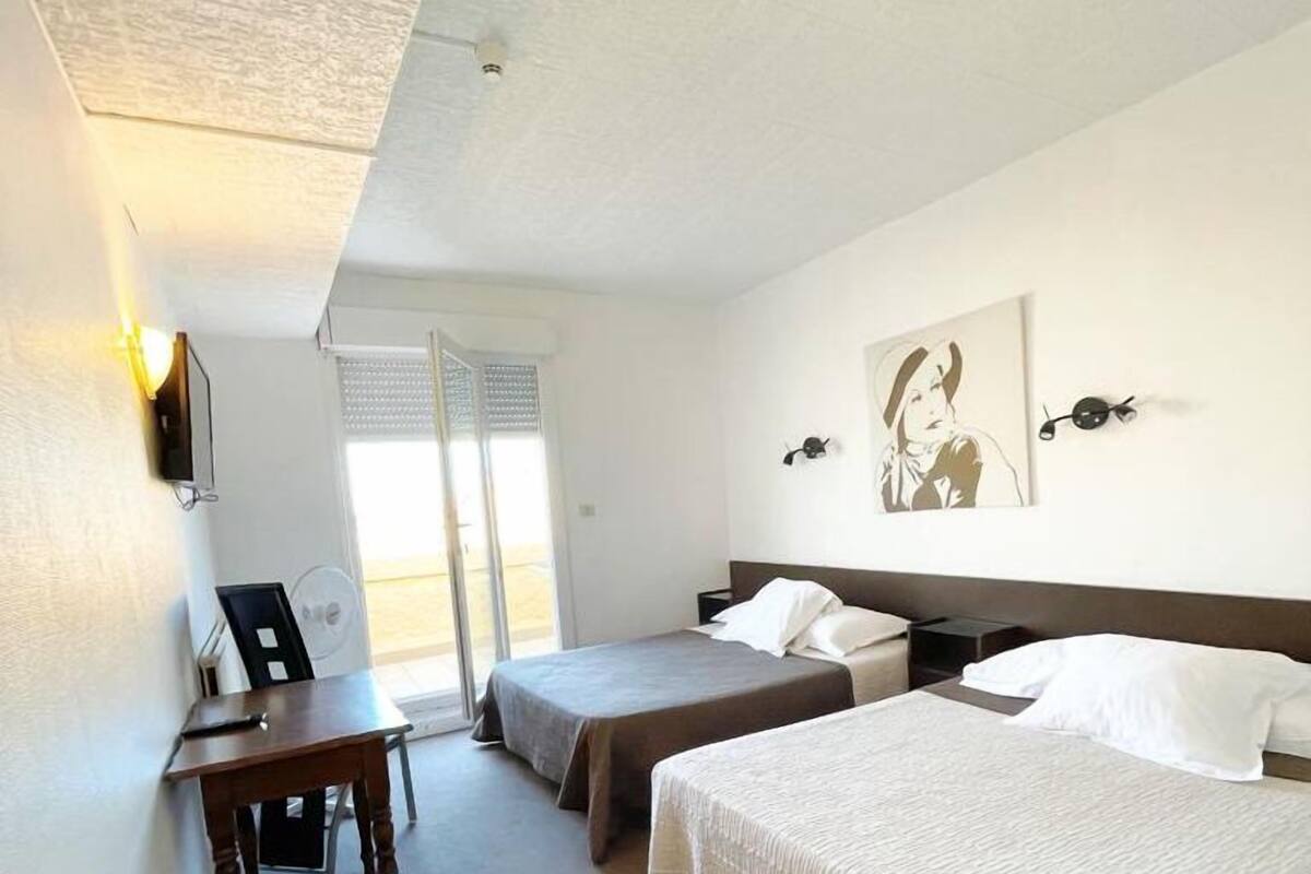 Une chambre d’hôtel avec deux lits simples, un bureau et une chaise. Il y a une fenêtre avec des stores, une horloge murale et un tableau encadré.