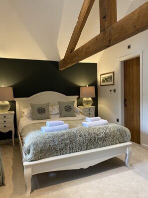 Quarto com cama branca, duas mesas de cabeceira, parede escura, vigamento de madeira no teto e uma porta.