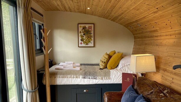 A cozy room with a wooden ceiling, a bed with pillows, a wooden coat rack, a desk with a lamp, and a window with curtains. - Robin’s Nest - ( Top Cabin )  - sleeps 2 guests (Foolow)