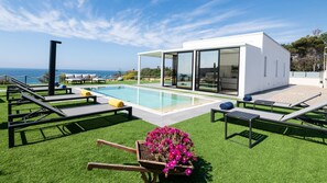 A modern outdoor pool area with lounge chairs, a small garden, and a building with large glass doors. - Villa Sabbie Rosse 6 2 (Isola di Capo Rizzuto)