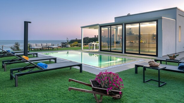 A modern outdoor pool area with lounge chairs, a dining setup, and a view of the ocean. - Villa Sabbie Rosse 6 2 (Isola di Capo Rizzuto)