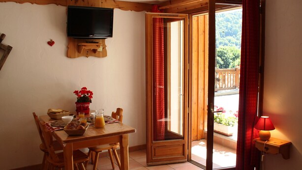 A dining area with a wooden table, chairs, a television mounted on the wall, and a view of the outdoors through an open door. - Studio cabin Valloire 4 pers South - Pets allowed, Wifi (Valloire)