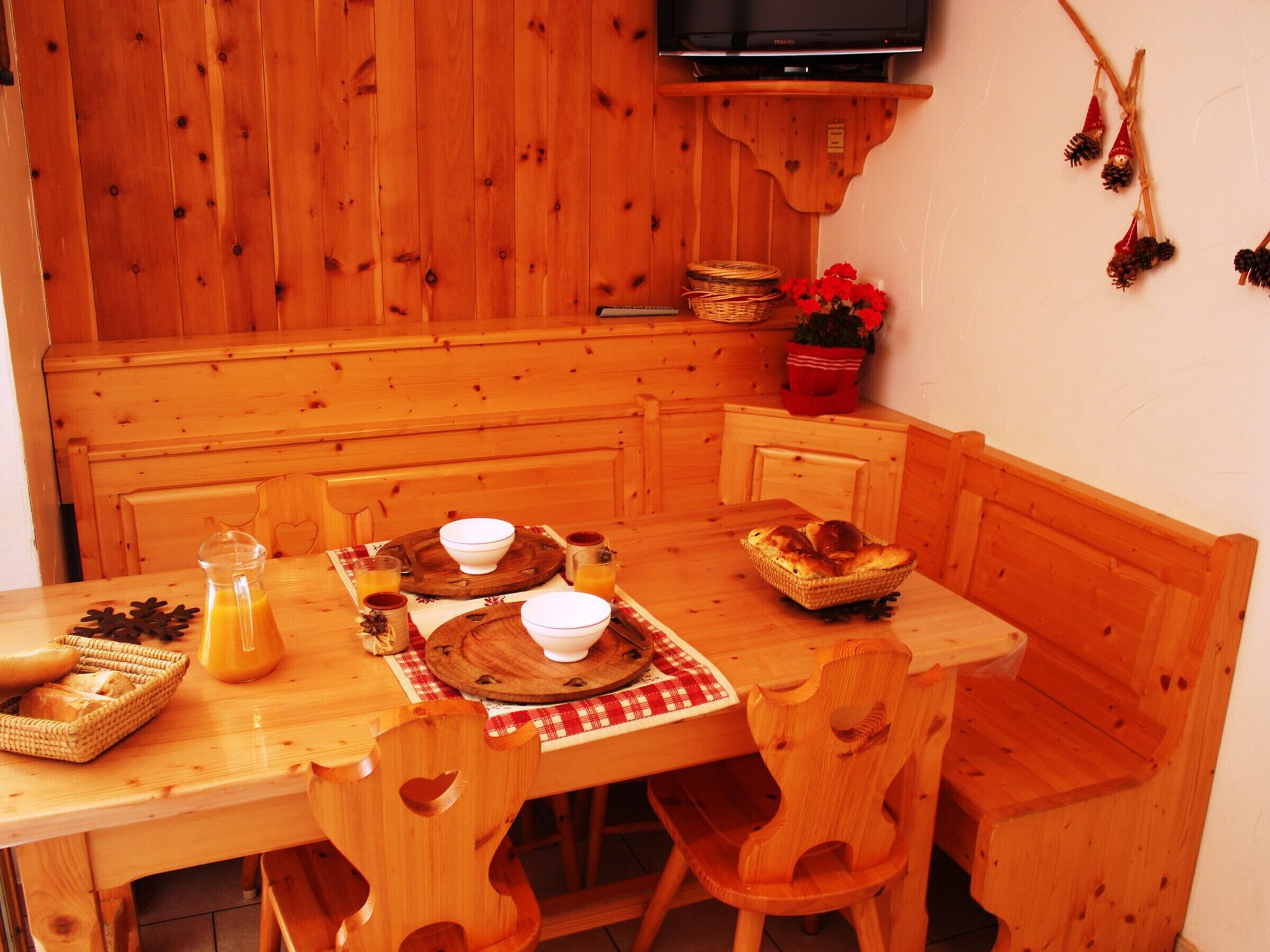 A wooden dining table with chairs, a television above, and a kitchen area with wooden cabinets and a red flower pot.