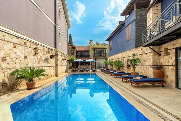 Una piscina con sillas de descanso y plantas en macetas, rodeada de muros de piedra y un cielo azul despejado.