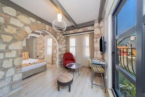 Una habitación con pared de piedra, una cama, una silla roja, una mesita y vistas a través de un ventanal.