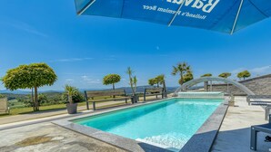 Une piscine offrant une vue dégagée sur le paysage environnant, une pergola surmontée d’une toile bleue et un mur en pierre.