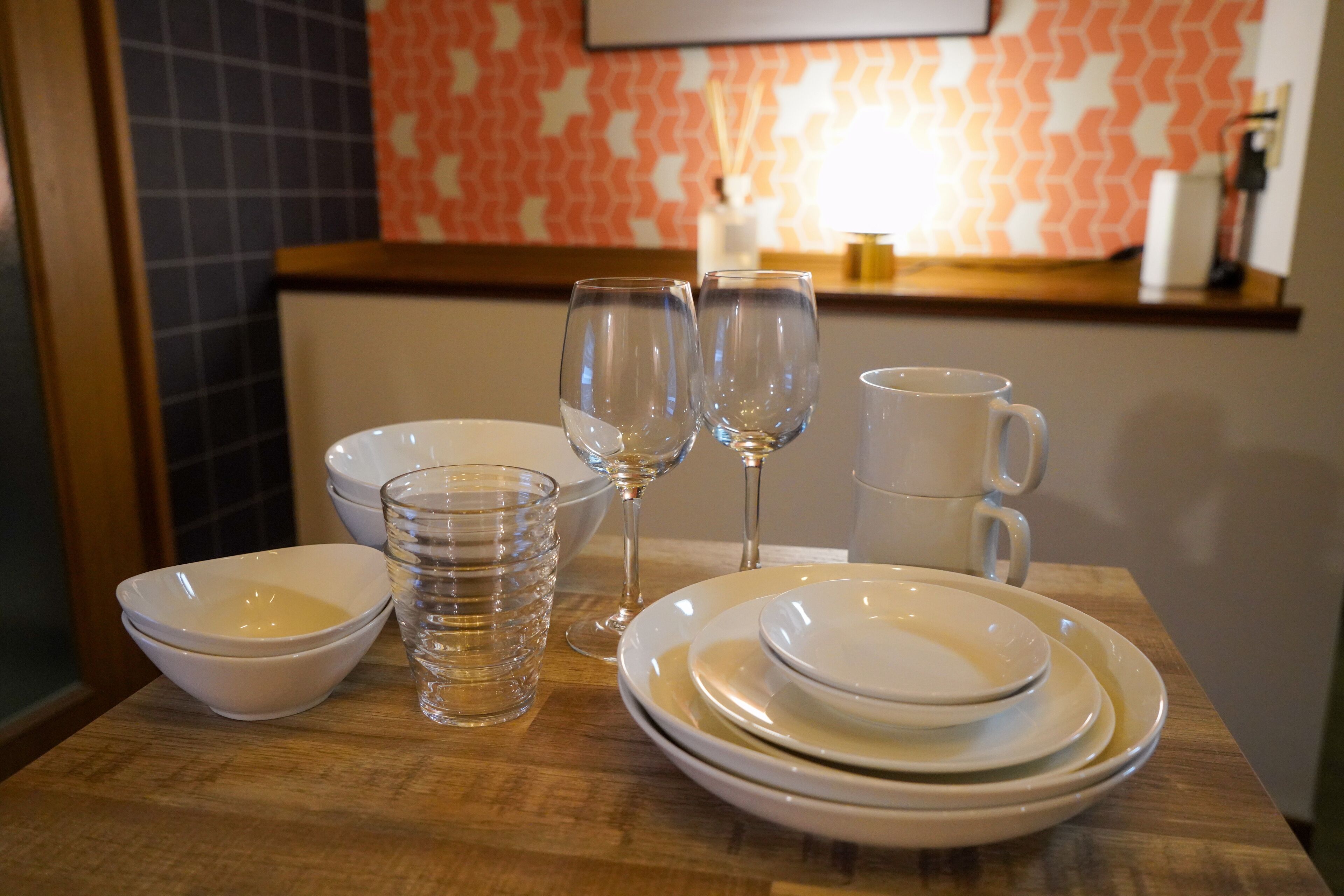 Una mesa de comedor dispuesta con platos blancos, tazones y vasos transparentes, con una pared estampada y una estantería al fondo.