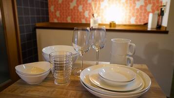 Una mesa de comedor dispuesta con platos blancos, tazones y vasos transparentes, con una pared estampada y una estantería al fondo.