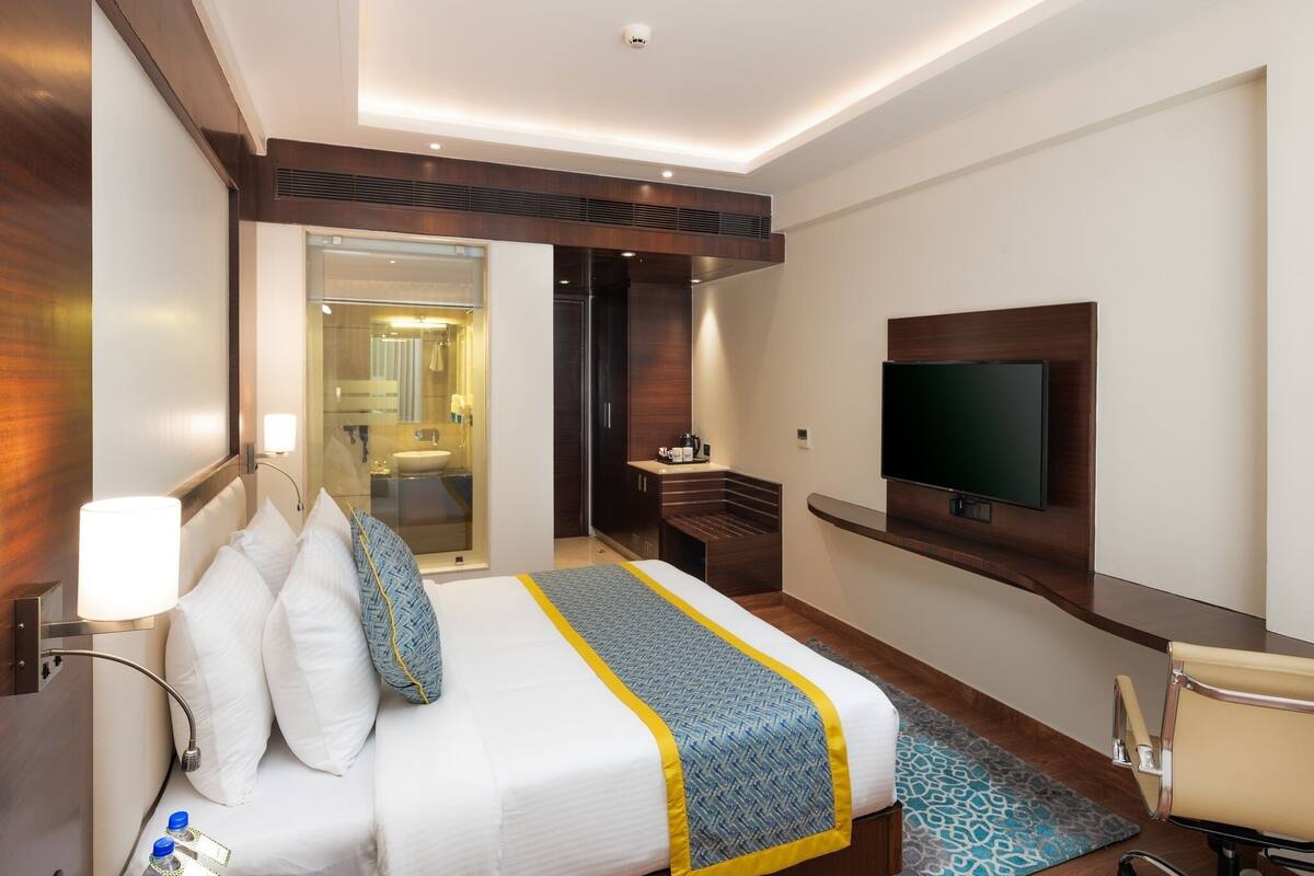 Habitación de hotel moderna con una cama grande, un televisor de pantalla plana montado en la pared, un escritorio con lámpara, y un baño visible a través de una puerta abierta.