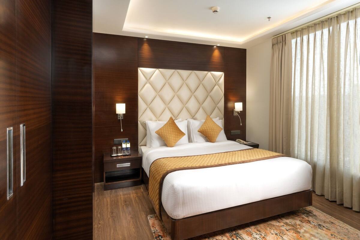 Habitación de hotel con una cama grande, paneles de madera, un cabecero capitonado y vistas de la ciudad a través de cortinas translúcidas.