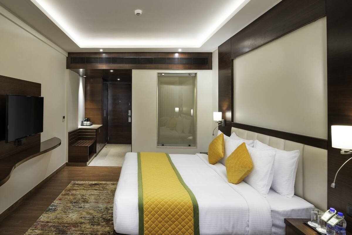 Habitación de hotel con cama, mesita de noche, televisor y baño con ducha de vidrio.