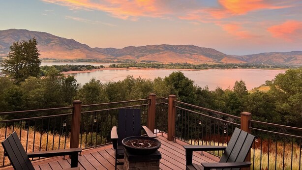 Une terrasse avec coin salon extérieur et foyer, offrant une vue sur un lac et des montagnes.