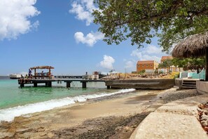 A beach with a pier, a thatched-roof structure, and a building with a red roof. - Chachacha Apartment At Sea with spectacular views and dining with an ocean view (Kralendijk)