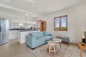 A modern kitchen with white cabinets, a blue sofa, a round coffee table, and a view of greenery through a window.