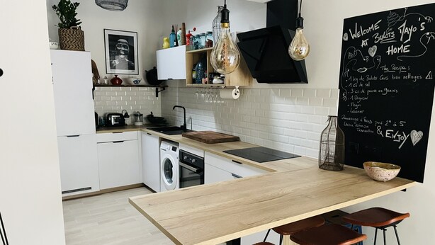 A modern kitchen with a wooden table, bar stools, a blackboard with recipes, and a washing machine. - Loft Chaleureux à 200 Mètres de la Plage (Wimereux)