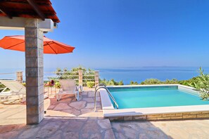 Una piscina con vista despejada al mar, un patio de piedra y un paraguas rojo.