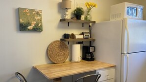 A compact kitchen with a wooden table, two black chairs, a refrigerator, a microwave, a coffee maker, a lamp, a potted plant, a vase with yellow flowers, a woven basket, and a painting of flowers on the wall. - Cozy Central Studio - Gunnison (Gunnison)