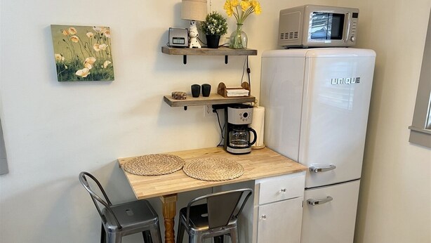 A compact kitchenette with a white refrigerator, a coffee maker, and a microwave on top. There is a wooden shelf above the counter holding a clock, a small potted plant, and a vase with yellow flowers. Two woven placemats are on the counter, and there are two black chairs. - Cozy Central Studio - Gunnison (Gunnison)