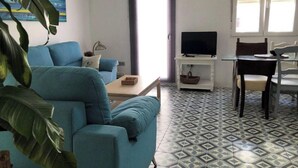 Un salon avec un canapé bleu, un téléviseur posé sur un meuble blanc, une table à manger avec des chaises et des carreaux de sol à motifs.